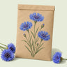 Kraft seed packet illustrated with Blue Boy Bachelor's Button, with fresh Blue Boy Bachelor's Button beside it